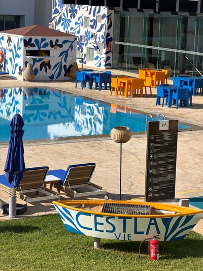 The image depicts an outdoor pool area in Nador, Morocco. A swimming pool with blue and white floral tile accents runs through the scene. Tables and chairs in blue and orange hues are positioned along the pool's edge. A sign displaying pool regulations in Arabic and French stands near the pool. Lounge chairs with blue cushions are located next to the pool, near a blue umbrella. A decorative surfboard with the text "C'EST LA VIE" and a fire extinguisher attached is placed on the grassy area adjacent to the pool. A building with blue and white floral designs is visible in the background. The overall environment suggests a resort or hotel setting.