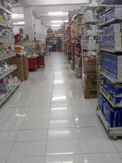 The image displays an interior aisle of a retail establishment located in Nganjuk, Indonesia. The floor is composed of reflective white ceramic tiles with dark grout lines, extending directly from the foreground into the background. Overhead, multiple fluorescent strip lights illuminate the space, their reflections clearly visible on the polished floor. White structural columns are present, supporting the ceiling.
On the left side of the aisle, a multi-tiered white metal shelving unit is stocked with various products, including items in clear plastic packaging, bags of orange-colored goods, and stacked cardboard boxes. Further down this left aisle, clothing items, specifically what appear to be baby garments, are displayed on hangers, adjacent to two red mobile display tables.
The right side of the aisle features another extensive multi-tiered white metal shelving unit, densely filled with a wide array of consumer goods in boxes and bags. Visible packaging includes blue-wrapped items and brown cardboard boxes, one of which partially displays the text "SUN Baby Po...", indicating baby products. Numerous brown cardboard stock boxes are stacked on and beneath these shelves.
In the mid-ground, towards the distant end of the aisle, two individuals are observed. One individual, seen from behind, wears dark clothing and stands near a metal shopping cart. A second individual, wearing lighter attire, is partially visible nearby. The background reveals additional shelving units and merchandise.
