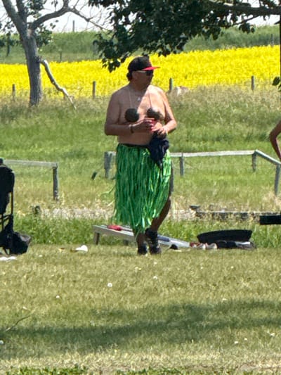 A person wearing a black baseball cap with a red brim and sunglasses is standing in a grassy area. They are shirtless and wearing a green grass skirt. They are holding two round objects in their hands. In the background, there is a field of yellow flowers and trees. The ground is covered in green grass with scattered white flowers and some fallen leaves. There are also some wooden posts and fences visible in the background. The image appears to be taken outdoors in a rural or park-like setting. The location is not specified in the image.