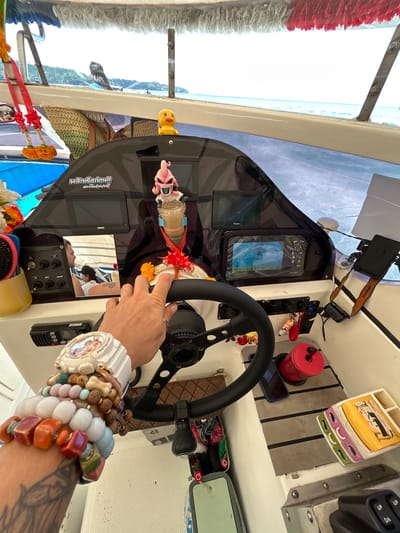 The image depicts the interior of a boat, likely a speed boat, on the water near Ko, Thailand. The foreground is dominated by a person's arm, adorned with numerous colorful bracelets and a white watch, resting on the steering wheel. The arm is tattooed with what appears to be floral designs.
The boat's dashboard is visible, featuring several electronic displays, including a GPS unit showing navigation data. A small, pink figurine resembling Majin Buu from Dragon Ball Z is placed on the dashboard, alongside a yellow duck figurine. Two red ribbons with orange flowers are tied to the boat's structure. A photograph of two individuals is also visible on the dashboard.
Other objects on the boat include a red cylindrical container, a dark-colored smartphone, a black radio or communication device, and a compartmentalized tray with what looks like small figures or images. In the background, the calm blue sea and a lush green island coastline are visible, characteristic of the tropical scenery in Thailand. The overall impression is a snapshot of a leisurely boat trip in a scenic, tropical setting.