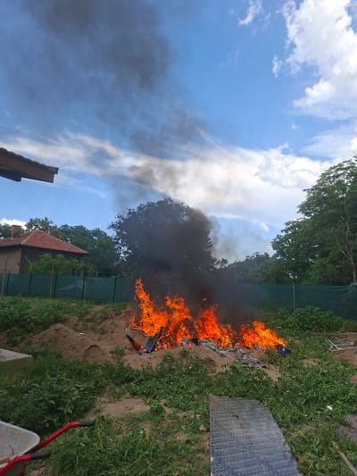 The media file depicts an outdoor scene with a fire burning in the middle of a sandy area, surrounded by lush green vegetation. The fire is intense, with bright orange flames reaching upwards, and a thick plume of dark smoke billows into the sky. In the background, trees and a house with a red-tiled roof are visible. A green fence runs along the edge of the property. To the right of the fire, a large piece of dark material, possibly plywood or a sheet of metal, rests on the ground. A red-handled wheelbarrow is also visible in the lower right corner.
The scene appears to be a backyard or garden area in Dolna Malina, Bulgaria. The bright sunlight and blue sky with scattered clouds suggest it is daytime, likely late morning or early afternoon. The presence of fire and smoke indicates an outdoor burning event, possibly of trash or debris. No people are visible in the frame.
Notable details include the vibrant flames, the contrast between the burning fire and the green foliage, and the dark smoke rising against the blue sky. The overall impression is one of an active burning process in a semi-rural or residential setting. There is no visible text in the image.