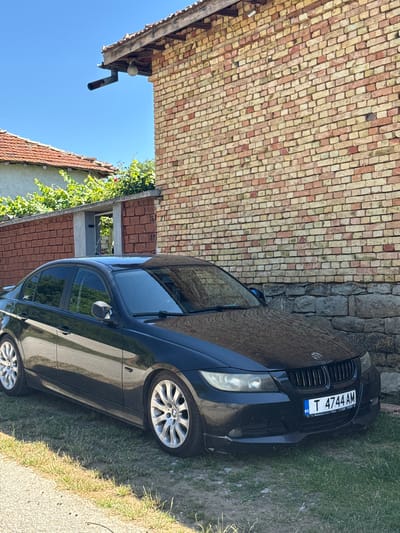 A black BMW sedan is parked on grass in front of a brick building in Konop, Bulgaria. The building has a red-tiled roof and a rough stone foundation. The car's license plate is T 4744 AM. The sky is clear and blue.
