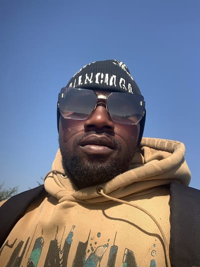 A close-up, portrait-style, vertical shot shows a man wearing a black beanie with white lettering and a tan hooded sweatshirt. He is also wearing large, dark sunglasses. The background is a clear blue sky. The lighting suggests it is daytime, likely a sunny day. The man has a beard and appears to be looking directly at the camera. The photograph seems to be taken outdoors. Some of the white lettering on the beanie is visible, possibly reading "NYC" or a similar phrase. The hooded sweatshirt has a graphic design on it, with abstract shapes and lines in black, blue, and teal. The overall mood of the image is casual and possibly confident. No specific activity or event is clearly depicted, other than the man posing for a photo.