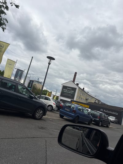The image shows a street scene in Bielefeld, Germany, marked by the presence of a car dealership, "AS Automobile". Several vehicles are parked along the roadside, including a blue Opel Corsa with the license plate "BI E EV 88", and a black SUV. The view is partially obscured by a side mirror of a vehicle, giving the impression of being photographed from inside a car. Flags bearing the Opel logo are visible, along with a sign for "Europcar" advertising car and truck rentals at "Eckendorfer Strasse 34". The sky is overcast with heavy clouds.