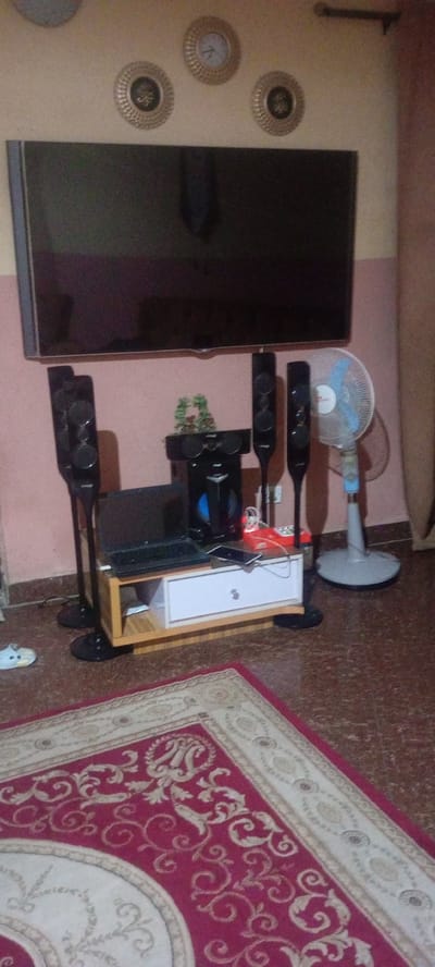 This image captures a home entertainment setup within an indoor living space, located in Rijiyar Zaki, Nigeria. The central feature is a large, dark-screened flat-screen television mounted on a warm pink-colored wall.

Below the television, a home theater system is arranged. This includes a central unit with a visible blue light, positioned on a light wood and white media stand. Four tall, slender black speakers stand on the floor, two on either side of the media stand. A black laptop is open on the left side of the stand, and a dark-colored smartphone rests on the right. To the far right of this setup, a light blue and white standing fan is visible.

The floor is covered with brown speckled tiles, with a large, ornate red and cream-patterned rug extending across the foreground. A single light-colored piece of footwear is on the tiled floor to the left. Above the television, three circular, gold-colored wall decorations are mounted; one appears to be a small clock, and the other two feature what resembles Arabic calligraphy. The scene is static, showing a tidy interior with no people or specific activities taking place. The lighting suggests an indoor environment.