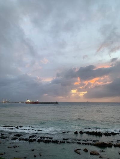 The image displays a coastal scene in Ksar El Majaz, Morocco, at either dawn or dusk, characterized by a predominantly overcast sky. The sky is filled with dark gray and lighter clouds, with distinct patches of orange and yellow light emanating from the mid-right. The immediate foreground features a rocky shoreline with dark, irregular rock formations partially submerged in the sea. Waves are breaking over these rocks, creating white foam. The sea's surface, a muted blue-gray, extends towards the horizon. In the mid-ground to the left, a substantial concrete pier or breakwater projects into the water, equipped with multiple active light sources. A large cargo ship with a red hull is positioned near this pier. A smaller, darker vessel is faintly visible on the distant horizon to the right. A small lighthouse structure is located at the far end of the prominent pier. Several small birds are observed in flight over the water. No human subjects are discernible.