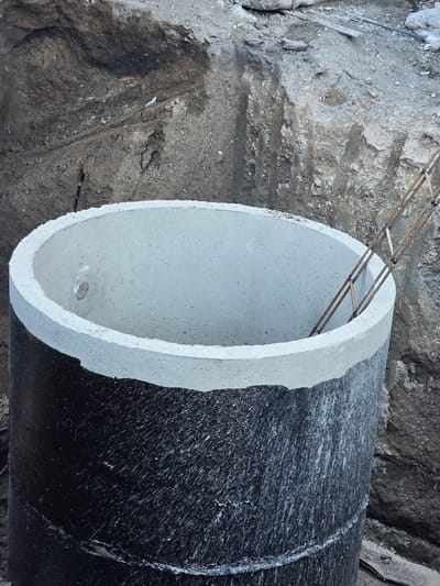 This is a medium-shot, close-up, eye-level, outdoor, daytime photograph of a large, concrete pipe section at a construction site in Perugia, Italy. The pipe is black on the bottom half and white on the top half, with a beveled edge where the colors meet. The interior of the pipe is visible and appears to be made of concrete. Inside the pipe, a section of metal rebar cage is visible. The pipe is situated in a dug-out area with dark soil and rocks surrounding it. The soil shows signs of excavation and has a rough, uneven texture. The lighting is natural and casts soft shadows. The image is in focus and the colors are natural. There are no people visible in the image. The overall impression is one of ongoing construction work.