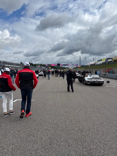 The image depicts an outdoor scene on an asphalt race track in Hohenstein-Ernstthal, Germany. The sky is predominantly overcast with scattered clouds. Numerous individuals are walking on the track surface, which features white painted markings. In the foreground, a male and a female are walking away from the camera; both wear red and black jackets with 'PORSCHE' branding and white baseball caps. To the right, a white and black prototype-style race car, branded with 'MOBIL 1' and 'HYPERCAR', is stationary with its driver-side door open, while a person stands beside it. Additional race cars and a large crowd of people are visible further down the track. The background includes track barriers, grandstands populated by spectators, event tents, and large banners displaying 'BWT', 'Change World', 'VAICO', and 'PORSCHE' logos. A digital display screen in the background shows '00:14:54'.