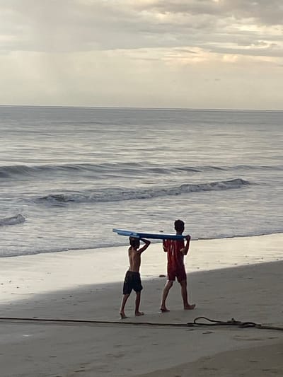 Two young males are walking on a sandy beach in Bateetimoh, Indonesia. The male on the left is wearing dark shorts and is carrying a blue boogie board over his head. The male on the right is wearing red shorts and a red t-shirt with white stripes. He is also carrying the boogie board over his head. They are walking towards the right of the frame. The ocean is to their left, with gentle waves breaking on the shore. The sky is overcast with a light haze. The sand is wet and reflects the light from the sky.