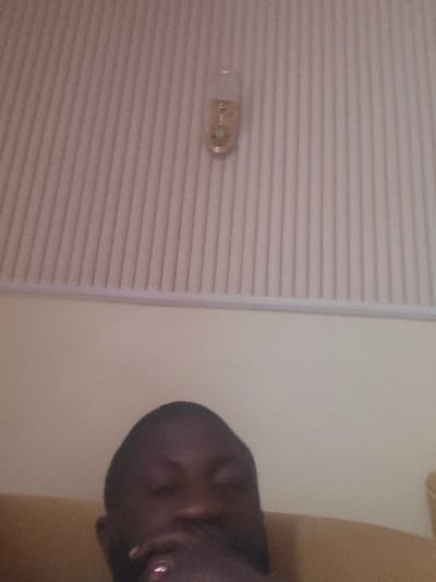 The scene depicts an interior location in Babawa, Nigeria. An adult male, appearing to be of dark complexion, occupies the lower portion of the frame, presented from the neck up. His eyes are closed or heavily lidded. His right hand is positioned near his mouth, with a reddish object, possibly a ring or painted nail, visible on one finger. He is reclining against a light brown, possibly upholstered, surface. The upper half of the image displays a light-colored wall or ceiling characterized by distinct vertical ridges. Affixed to this surface in the upper right quadrant is a translucent, elongated glass object, possibly a decorative light or an air freshener unit, containing a pale yellow liquid and a small greenish insert. No direct interactions or specific actions beyond reclining are discernible.
