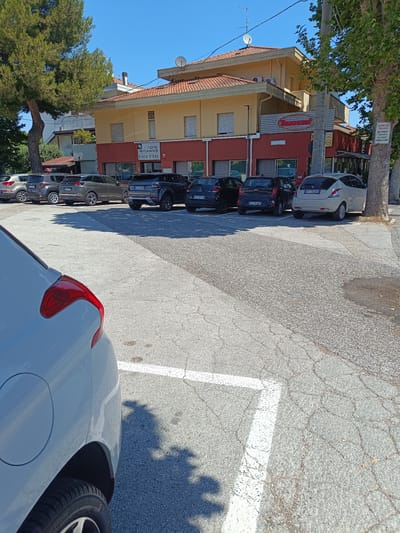 The image shows a parking lot in front of a building that houses a hotel and a restaurant. The building is yellow with a red lower section and a red-tiled roof. A sign on the building reads "HOTEL RISTORANTE PIADA D'ORO". There are several cars parked in the lot, including a white Fiat 500, a black Kia, a grey Peugeot, and a dark grey Range Rover. The parking lot is paved with asphalt and has white parking lines. A street sign attached to a tree on the right side of the image reads "PROPRIETA' PRIVATA Parcheggio riservato ai clienti PIADA D'ORO". The sky is clear and blue. The overall scene depicts a typical day in a commercial area. The location is Riccione, Italy, as indicated by the street sign.