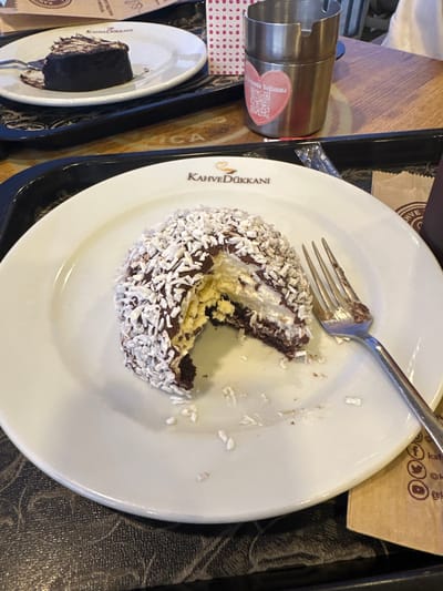 The image shows a partially eaten dessert on a white plate with the logo "Kahve Dükanı" in a cafe setting. A fork rests on the plate near the dessert, which is a round pastry covered in white coconut flakes, with layers of chocolate cake and a cream or custard filling visible. The plate sits on a black tray, along with a metal container, a small plate with another piece of chocolate cake, and other items. The background shows glimpses of a table surface and what appears to be a marketing material. The location is Köyceğiz, Turkey, based on the cafe's name, "Kahve Dükanı," which is a common Turkish coffee shop chain.