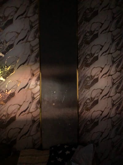 The image shows a surface with a marble-like pattern in shades of gray, white, and brown with dark veining. A dark gray, possibly metallic, rectangular object is positioned horizontally in the center of the frame, appearing to be bordered by gold trim. In the top right corner, a portion of a pillow or cushion with a star pattern is visible. The lighting is dim, casting shadows across the scene. The image may be in Rantopanjang, Indonesia, but there are no elements in the picture that can confirm the city as the source of the picture.