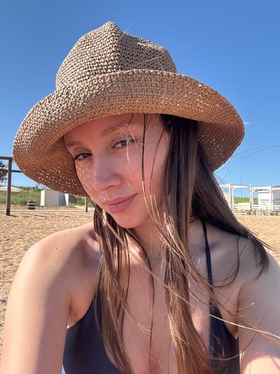 A young woman is posing for a close-up selfie at a beach location in Береговой сельский совет, Ukraine. She is wearing a straw hat that partially obscures her face with shadows. The sky is clear and blue, visible in the background. In the distance, there are structures that could be part of a beach resort or establishment. The woman is wearing a dark-colored swimsuit. Her hair is long and brown, with strands blowing in the wind.