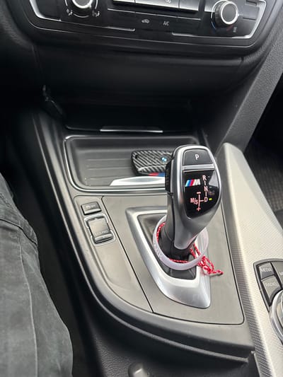 The image depicts the interior of a BMW vehicle, specifically the center console and lower dashboard area, viewed from the driver's perspective in Suhache, Bulgaria. The automatic gear selector, currently in "P" (Park) mode, is prominently featured, displaying the BMW M logo and shift options for R, N, D, and M/S. A white plastic ring marked "SPARK" encircles the base of the gear selector, from which a red and white braided string hangs. To the left of the gear selector are three buttons: "OFF" (likely for traction control), "SPORT", and "ECO PRO". Above the gear selector, within an open storage compartment, rests a BMW key fob with a carbon fiber pattern cover and BMW M colored stripe detailing. Parts of the climate control unit, including knobs and "A/C" button, are visible in the upper background. On the far left, the knee and upper thigh of an individual wearing dark trousers are visible, indicating a person is seated in the driver's position. The interior features black and grey finishes, including textured plastic surfaces.