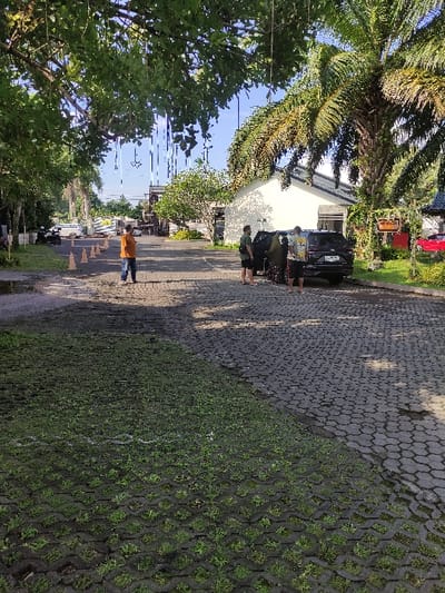 The image shows a street in Gambiran, Indonesia, located at coordinates -8.434 latitude and 114.18 longitude. The street is paved with gray interlocking pavers. The right side of the street is lined with green grass growing in the spaces between the pavers. There is a small, dark-colored car parked on the left side of the street. Two people are walking away from the car toward the camera. A third person, wearing an orange shirt, is walking toward the car on the left side of the street. There are trees on both sides of the street. The sky is blue and the weather appears clear.