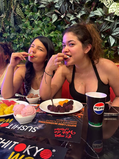 Two young women are captured in a close-up, top-down shot, seemingly enjoying a meal at a restaurant with a lively, green, plant-filled wall as a backdrop. Both women are casually dressed in tank tops. The woman in the foreground, on the left, has dark hair and is leaning towards the camera, holding a piece of food to her mouth. She appears to be smiling or laughing. Beside her, the second woman, with lighter brown hair, is also eating and looking towards the camera.
On the table in front of them are various food items, including bowls of what appears to be ice cream and fruit. A Pepsi cup is prominently visible, along with menus featuring text in Spanish. The menus display titles such as "El mejor Wok de Madrid," "BUFFET LIBRE INTERNACIONAL," and "SUSHITO." The overall atmosphere is casual and convivial, suggesting a shared meal or a fun outing. The lighting appears to be artificial, creating a vibrant and somewhat dramatic effect, with a purple hue visible on the left side of the image. The scene is set in a restaurant, with the text on the menus indicating a possible location in Madrid, Spain, and the mention of "Carretera De San Martín" potentially narrowing down the specific area.