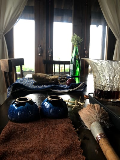 This is an indoor, eye-level shot of a wooden table set for a tea ceremony, likely in Thôn 4, Vietnam, given the natural light and rustic wooden architecture. In the foreground, two deep blue ceramic tea cups sit on a brown, textured cloth. To the right, a glass pitcher with a distinctive textured surface holds dark liquid, possibly tea, and is placed on a black tray. A whisk-like brush with a wooden handle and light-colored bristles rests on the table, angled towards the lower right corner. Behind the cups, a blue and white patterned cloth is laid out, upon which rests a small, light brown pouch and a single reddish-purple fruit. A tall, green glass bottle with a small bouquet of light green foliage on top stands in the mid-ground. In the background, large windows reveal a softly lit outdoor environment, with wooden beams and architectural elements suggesting a traditional Vietnamese setting. The overall impression is one of a serene and artisanal tea ritual in a rustic, natural setting.