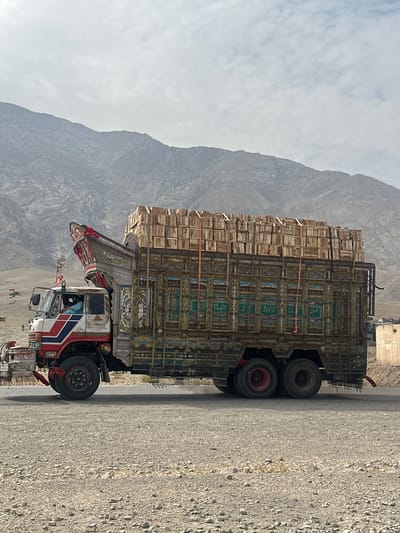 An eastbound, heavily laden, ornate truck is captured in profile, traveling on a paved road that cuts through a rugged, arid landscape under a hazy sky. The truck, a prominent subject, is adorned with vibrant colors and intricate designs, typical of Pakistani truck art. It is loaded with numerous wooden crates stacked high, secured by orange straps. A man, dressed in a light blue garment, is visible driving the truck. In the distance, behind the truck, a range of imposing, rocky mountains dominates the background. The foreground is a gravelly, dusty terrain. The road appears to be in a rural or semi-arid region, possibly along a transportation route in Pakistan, given the style of the truck. The overall impression is one of a journey through a challenging but visually striking environment.