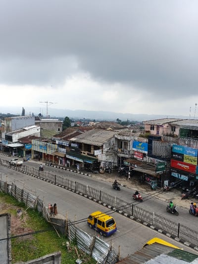The image depicts an elevated view of a city street in Kidapawan, Philippines, under a uniformly overcast, gray sky. The scene is dominated by a two-way asphalt road running diagonally through the midground, separated by concrete barriers topped with black metal railings.
Along the right side of the road, numerous commercial buildings of varying architectural styles and states of repair line the street. Many display prominent advertisements for mobile phone brands such as Oppo, Vivo, Infinix, Realme, and Ringtone, alongside signs for DasLig and other businesses. Multiple motorcycles are parked along this sidewalk or in motion on the road. Specifically, a black motorcycle with a rider is visible in the right lane, followed by a red motorcycle with two riders, and further right, a green motorcycle with a rider, and a blue motorcycle with two riders.
On the left side of the road, more commercial structures are visible, some with awnings. A white pickup truck is parked on the far left. On the road itself, a distinctive yellow van with blue lower trim is positioned in the left lane, moving away from the viewer. A small group of three individuals, one appearing to wear a head covering, stands on a raised grassy embankment to the left of the road, partially enclosed by a makeshift fence of corrugated metal sheets.
In the background, distant hazy mountains or hills are visible beneath the heavy cloud cover, framing the urban landscape. A tall, multi-level antenna tower is visible on the left skyline, rising above the buildings. The foreground on the left shows a concrete structure and green vegetation, partially obscured by the image frame. The scene indicates active urban commerce and transit.