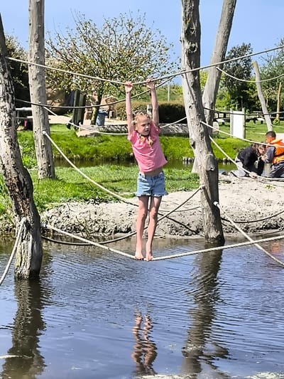 A young girl wearing a pink shirt and denim shorts is walking barefoot on a rope bridge that crosses over a body of water. She is holding onto a higher rope with both hands for balance. The bridge is supported by wooden posts. The girl's reflection is visible in the water. In the background, there are other people, including some wearing orange life vests. The setting appears to be a park-like area in Sint Maartensvlotbrug, Netherlands.
