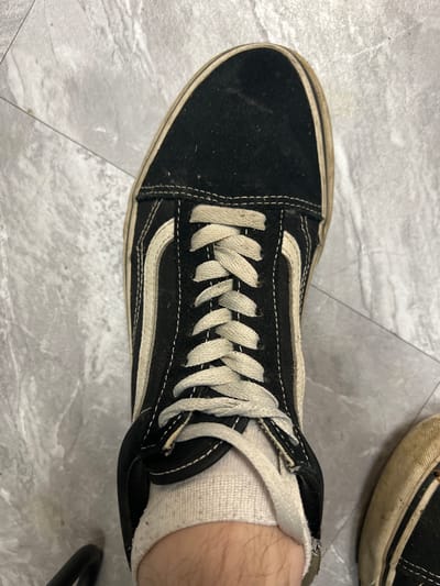 A high-angle, close-up shot shows a black Vans Old Skool sneaker with white laces and a white stripe on a textured gray tiled floor. The shoe is worn and slightly dirty, with visible wear on the sole and fabric. A portion of a white sock is visible around the ankle area, and the leg of the wearer is also visible. Another similar shoe is partially visible on the right side of the frame. The environment appears to be indoors, possibly a home or a public space with tiled flooring. There is no indication of the shoe's location in Ленинский, Russia.