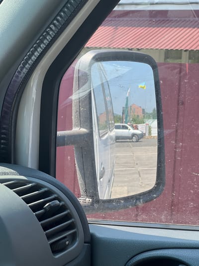 The image is taken from inside a vehicle, looking out the driver's side window. A side-view mirror is prominently featured, reflecting the external environment. The mirror shows a silver SUV driving on a paved surface, with a red-tiled roof structure and flags in the background. One of the flags appears to be the Ukrainian flag. The interior of the vehicle is visible in the foreground, including a dark grey dashboard with an air vent and part of the steering wheel. The exterior of the vehicle's door is visible through the window, and it is a dark red color. The overall scene appears to be an outdoor urban or semi-urban setting in Khust, Ukraine, likely during daylight hours given the brightness and shadows.