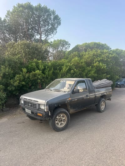 A dark gray Nissan pickup truck is parked on an asphalt road in a rural or semi-rural area. The truck has a single cab and is loaded in the bed with a bundle of long, thin wooden poles or branches tied together. The truck is dirty and appears somewhat weathered. In the background, there is a dense green hedge or shrubbery, and beyond that, tall pine trees under a clear blue sky. To the right of the pickup truck, a dark blue SUV is visible parked further down the road. The license plate on the pickup truck is partially visible and reads "CA 652495".