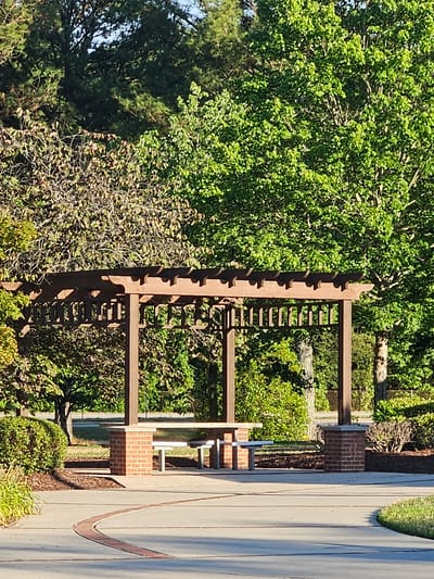In the city of Dortches, United States of America, a park scene unfolds with a wooden pergola as the central element, casting dappled shadows on the concrete pathway and picnic tables beneath it. The pergola is supported by brick pillars and features a slatted roof, suggesting it provides shade. Two white picnic tables, one with attached benches, are visible under the pergola. The surroundings are lush with greenery; a mix of deciduous trees with vibrant green leaves and trees with sparser, darker foliage, as well as manicured bushes and ground cover, create a natural backdrop. A paved pathway, curving through the scene, is highlighted by a decorative brick inlay. The lighting indicates it is daytime, likely late afternoon, with the sun casting long shadows and illuminating the foliage. No people are present in the image.