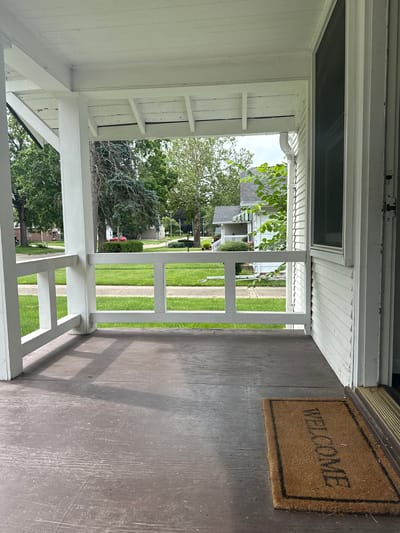 The image depicts the view from the porch of a house, looking out onto a suburban street. The porch itself is made of weathered wood planks and has white railings. A welcome mat is visible on the porch floor. Beyond the porch, a well-maintained lawn stretches to the street, with houses and trees lining the opposite side. The sky is overcast, suggesting a cloudy day. The scene appears to be a residential area in a city.