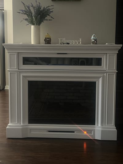 This is a head-on, medium shot of a white electric fireplace with a glass-fronted firebox, set against a neutral wall. The fireplace has a decorative mantelpiece on top, adorned with a white vase filled with artificial lavender, two small decorative figures resembling gnomes or whimsical houses, and a silver word art piece spelling "Blessed" with a key lying next to it. The firebox below the mantelpiece has a glass front revealing a brick interior and a dark floor. Below the glass, there is a control panel with a red indicator light illuminated, suggesting the fireplace is on. The fireplace sits on a dark wood-plank floor. The background is plain and light-colored, with a slight shadow cast on the left side, suggesting a light source from the left. There are no people visible in the image. The location of this image is not determinable from the visual information provided.