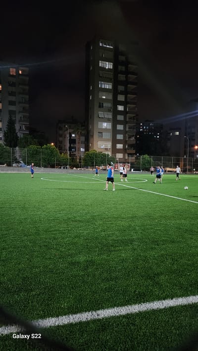 A group of young men are playing soccer on a brightly lit artificial turf field at night in a city. The field has white lines demarcating its boundaries and center circle. In the background, tall apartment buildings with illuminated windows are visible against the dark night sky. Trees and a fence are also present in the background, suggesting an urban park or recreational area. The men are wearing athletic clothing, with some in blue shirts and black shorts, and others in white shirts and black shorts. One man in the foreground is wearing a blue and red shirt. The overall scene depicts a casual game of soccer taking place in a city park. The location of the city is Çukurova, Turkey.