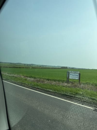The image captures a rural landscape near Thrapston, United Kingdom, viewed from the interior of a vehicle. A dark grey tarmac road with a distinct white edge line occupies the lower portion of the frame. Adjacent to the road, a narrow strip of dry grass with scattered white plant growth forms a verge, which transitions into a large expanse of vibrant green fields. In the right mid-ground, a freestanding wooden sign with white panels and black text is positioned within the field. The sign reads: "TITCHMARSH SHOP", "The WheatSheaf at Titchmarsh", and "PUB & RESTAURANT" with the phone number "01832 730261". The background features further green fields interspersed with distant hedgerows and a faint line of trees against a pale, hazy blue sky. No individuals or specific activities are present. The scene is illuminated by daylight.