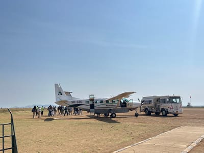 The image depicts an active airfield scene at Seronera, Tanzania, under a clear blue sky. A white twin-engine propeller aircraft, registration 5H-CDL, featuring "REGIONAL AIR" text and a green "A" logo, is centrally parked with its passenger doors open. Approximately 15 to 20 individuals are clustered around the left side of the aircraft, appearing to be in the process of boarding or disembarking. One person within the group wears a high-visibility vest. To the right of the aircraft, a white fuel tanker truck, with "PME FUELS" visible on its side, is positioned. The ground surface consists of dry, reddish-brown dirt and sparse, dry grass. A segment of a concrete or paved pathway is visible in the foreground, extending from the bottom right. Distant, low-lying hills are discernible on the horizon to the left. A red flag on a pole stands upright in the far right mid-ground.