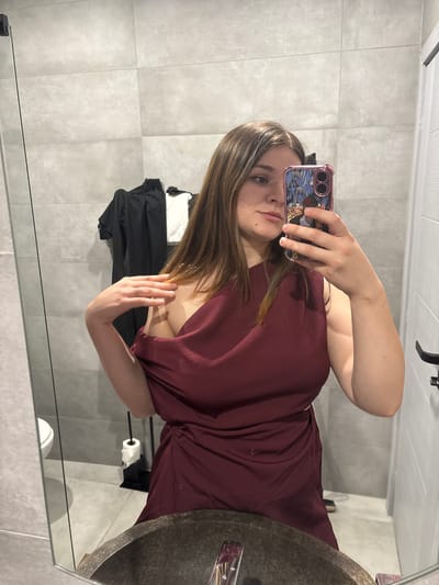 A young woman is taking a mirror selfie in a bathroom. She is wearing a dark red, one-shoulder dress with draping. Her right hand is resting on her shoulder, and she is holding a phone in her left hand, which has a cartoon character case. She has long brown hair and is wearing subtle makeup. The bathroom has grey tiled walls and a sink with a dark basin. A roll of toilet paper is visible in a holder behind her. Clothes are hanging on a rack behind her. The overall environment suggests a domestic setting within the city of Dolno Cherkovishte, Bulgaria.