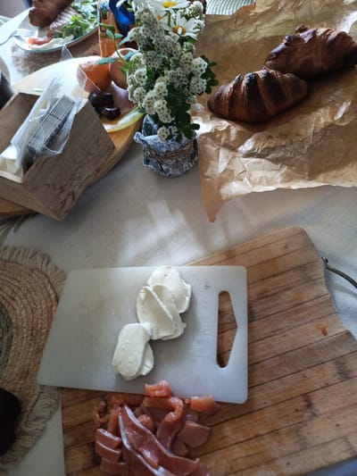 A close-up, overhead view captures a rustic breakfast spread on a light-colored tablecloth. In the foreground, a wooden cutting board holds a pile of chopped salmon and several slices of fresh mozzarella. Adjacent to it, a white cutting board with a cutout is also visible. To the upper left, a woven placemat with a fringed edge is partially in view, and behind it, a wooden box contains what appear to be packaged skewers or toothpicks. Further back, a bouquet of small white flowers in a patterned vase adds a touch of natural beauty. The background reveals two golden-brown croissants resting on crumpled brown paper. A glimpse of a plate with some food, possibly smoked salmon and greens, can be seen on the far left. Oranges and dark olives are also present on a small plate or surface. The scene suggests a leisurely breakfast prepared with fresh ingredients. There is no indication of the location in Alexandrovskaya, Russia.