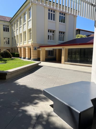 The image displays an outdoor architectural scene, likely a campus or institutional courtyard, under bright, clear sunlight in Kelvin Grove, Australia.
On the left side of the frame, a multi-story building with light-colored (cream or beige) walls is prominent, featuring numerous rectangular windows with white frames and distinct horizontal white architectural elements, possibly sunshades or decorative banding. A white slatted structure, resembling a pergola or awning, extends along the upper left edge of the image. The clear blue sky is visible above and to the left of the buildings.
To the right of the main building, a paved pathway made of light grey concrete slabs stretches into the background. Adjacent to this path, a narrow strip of vibrant green grass forms a garden bed, which contains a small, rounded bush. Further back, another building with a dark red-brown tiled roof and large glass-fronted sections is visible.
In the right foreground, a large, dark grey rectangular object, possibly a bench or a substantial planter box, sits beside the pathway. Strong sunlight casts well-defined, elongated shadows across the concrete pathways, including dappled patterns that suggest nearby trees or foliage.
No people are visible in the scene, creating a quiet and still atmosphere. A notable detail is a capital letter "A" displayed on a square sign attached to the cream-colored building, near what appears to be an entrance or transition area. The bright sun and sharp shadows indicate it is likely mid-morning or early afternoon.