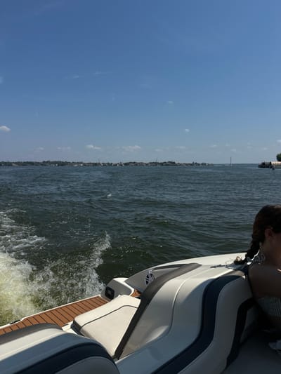 The image provides a stern-view perspective from a white and gray recreational boat moving on a body of water, located in the city of Montgomery, United States. The upper half of the frame shows a clear blue sky with sparse, wispy white clouds. The lower half features the water and the boat's stern.
The boat's stern section, equipped with brown synthetic decking and white upholstered seating with dark gray accents, occupies the lower foreground. A turbulent white wake trails directly behind the vessel in the dark blue-green water, indicating forward motion. The right side of a person's head and shoulder is partially visible in the lower right corner, seated within the boat. This individual has dark hair styled in two braids and is wearing a light-colored, possibly striped, top. A black speaker with a silver-colored emblem is mounted on the interior wall of the boat, visible near the person's shoulder.
The distant horizon reveals a continuous stretch of populated shoreline, densely packed with numerous residential-style buildings and green trees. Intermittent power transmission towers or similar tall structures are visible above the treeline along this distant landmass. On the far right side of the horizon, a distinct vertical structure, consistent with a bridge pylon or communication tower, stands over the water, adjacent to what appears to be a dock or other industrial-type structure extending from the shoreline.
