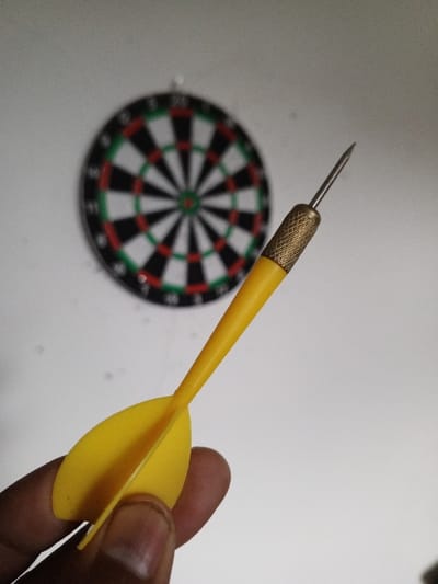 In Al Haouafate, Morocco, a close-up forensic image captures a human hand holding a dart in an indoor setting. The hand, visible with a thumb and index finger, secures the bright yellow-finned dart. The dart features a metallic, textured (knurled) gold-colored barrel section, transitioning into a pointed silver-colored steel tip. The yellow plastic flights of the dart are clearly visible. In the background, a circular dartboard, displaying traditional black, white, red, and green segments, is mounted on a plain white wall. The dartboard is out of focus, with the dart and hand in sharp foreground focus.