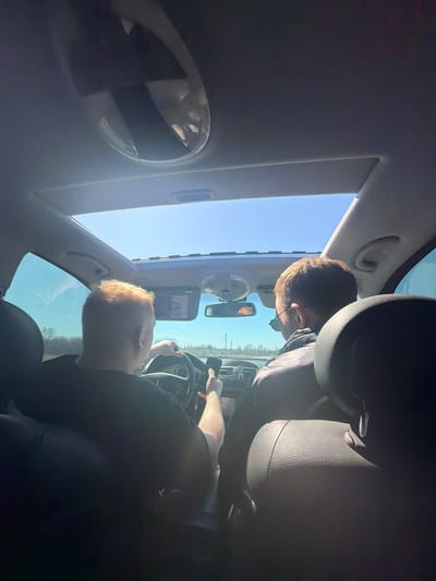 A low-angle photograph taken from within a car shows two individuals. One person is driving, their hands on the steering wheel. Another person is in the passenger seat. The car's sunroof is open, revealing a clear blue sky. A rearview mirror is visible. The location is inferred to be Гремяча, Russia based on user query.