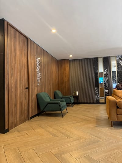 The image captures a portion of the interior of a business or hotel lounge in Boca del Río, Mexico. The design features a modern aesthetic with a blend of wood and dark stone-like surfaces. Two green chairs with black metal frames sit on a raised wooden platform, accompanied by small, clear side tables. A clear sign with text is affixed to the platform in front of the chairs. Above this area is a wall adorned with horizontal, textured panels and sections of black and white marbled material. Above these panels, a glimpse of a light brown leather sofa can be seen. The floor is tiled with light-colored wood in a herringbone pattern. The wall to the left is painted white. There are two recessed lights on the wall.