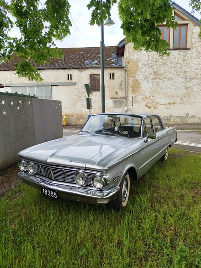 The image captures a classic silver car, identified as a Mercury Comet by the visible badge on its front fender, parked on green grass alongside a paved road in Manternach, Luxembourg. The car is presented from a front three-quarter view, showcasing its sleek design, prominent chrome grille, and quad headlights. It features chrome bumpers, window trim, and classic whitewall tires. A black license plate with white numbers "18355" is affixed to the front.

In the background, an old, light-colored building with a textured exterior, possibly stucco or roughcast, stands. The building shows signs of age and weathering, with some discolored patches on its walls. Wooden-framed windows are visible on the upper section of the building, and it has a tiled roof. A street light pole is positioned between the car and the building. Lush green trees with dense foliage frame the left side of the scene. The sky is overcast, suggesting daylight conditions without direct sunlight. The overall setting appears tranquil, depicting a vintage vehicle in a historic or rural European environment. No people are visible in the scene.