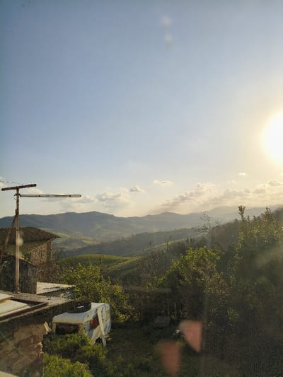 The image displays a rural landscape in Villa di Gemmano, Italy, under late afternoon lighting. The sky is clear blue with scattered white cumulus clouds, particularly dense along the distant horizon. A bright, low-angle sun is visible in the upper right, casting a warm light and producing noticeable lens flare.
In the left midground, the upper section of a rustic, light-colored stone building with a red-tiled roof is visible. An older model metallic television antenna is attached to a vertical pole, extending above this structure. Below and partially in front of the building, a white camper trailer with red markings, appearing disused or damaged, is situated on a grassy slope amidst dense green vegetation.
The background features multiple layers of undulating green hills and valleys, showing a mixture of forested sections and cultivated fields, receding into a light atmospheric haze towards the far horizon. No individuals are present, and no specific actions or interactions are depicted within the scene.