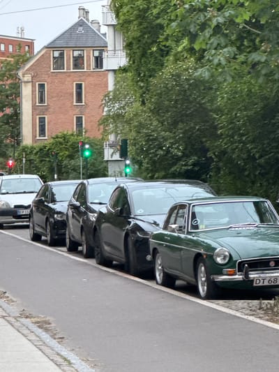In Frederiksberg, Denmark, a street scene is captured with several cars parked along the side of the road. The closest vehicle is a vintage green MG, which has the license plate "DT 68". Behind it, a row of other cars, mostly black sedans, are visible. A white car is parked further down the line on the left. Traffic lights are illuminated, with green lights showing for two of them and red for one. The background features a brick building and lush green trees, suggesting a residential or urban setting with foliage. The street appears to be paved with asphalt, and there's a sidewalk or pedestrian path made of concrete and brick. The sky is overcast.