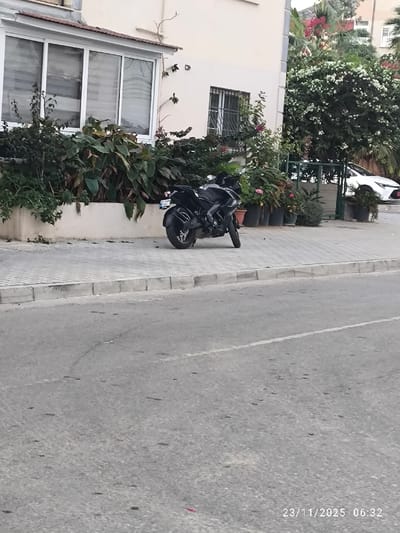 A black motorcycle is parked on the sidewalk in front of a building with lush greenery. Potted plants line the walkway. A white car is visible in the background. The street is in the foreground, occupying a significant portion of the frame. The photo appears to have been taken from a slightly low angle. The date stamp at the bottom right corner reads "23/11/2025 06:32."
