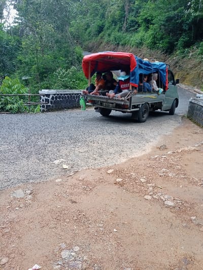 The image shows a grey pick-up truck with a red and blue canopy, traveling on a gravel road. The truck is traveling from left to right and is full of passengers. There are six passengers seated in the bed of the truck, facing forward. There is also a small green bottle on the left side of the image, near the truck. The truck is traveling on a paved road in a rural area, with a stone wall and lush vegetation on the left side. The road is flanked by dense foliage.  The location is in the town of Cisewu, Indonesia, which is located at latitude: -7.411, longitude: 107.542.  The image is taken from the perspective of a pedestrian who is standing to the left of the road, watching the truck travel away from them.