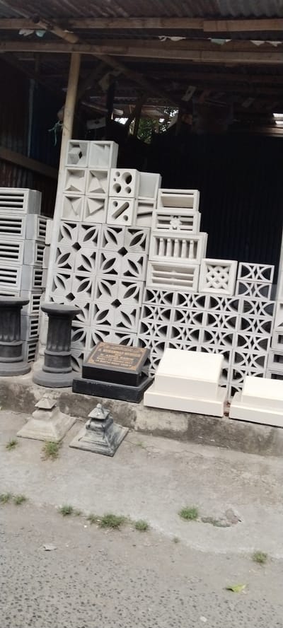 A collection of carved concrete blocks, likely decorative screens or ventilation bricks, are stacked and arranged outdoors under a weathered, corrugated metal roof supported by wooden beams. The blocks display various geometric and floral patterns. In the foreground, there are several stone pedestals of different sizes and designs, along with two dark, rectangular grave markers. One of these grave markers has an inscription in gold lettering. The ground is paved with concrete, with patches of sparse grass growing in cracks. The scene appears to be in a storage area or a workshop. The image may have been taken in the city of Mataram, Indonesia, given the context of Islamic grave markers. There are no people visible in the image.