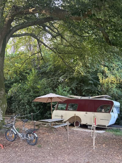 This image captures a tranquil outdoor scene, likely a campsite, nestled within a lush green environment characteristic of Saint-Vaast-du-Val, France. A vintage caravan, predominantly cream-colored with a deep red roof and accents, is parked on a bed of fallen leaves and mulch. The caravan's design suggests it is from an earlier era, featuring curved windows and a polished exterior.

In the foreground, a silver folding bicycle with front and rear baskets is parked, its wheels resting on the leaf-covered ground. A picnic table made of weathered wood, accompanied by an open, light-colored umbrella, is set up near the caravan, suggesting a space for relaxation and dining. The area is delineated by simple rope barriers supported by wooden posts.

Behind the caravan and picnic table, dense foliage and trees form a natural backdrop, with sunlight filtering through the leaves, creating dappled light and shadow. The overall impression is one of a peaceful, rustic retreat, bathed in the soft light of late afternoon or early morning. No people are visible in the frame, emphasizing the solitude and quietude of the location. The environment is natural and unmanicured, with a sense of undisturbed natural beauty.