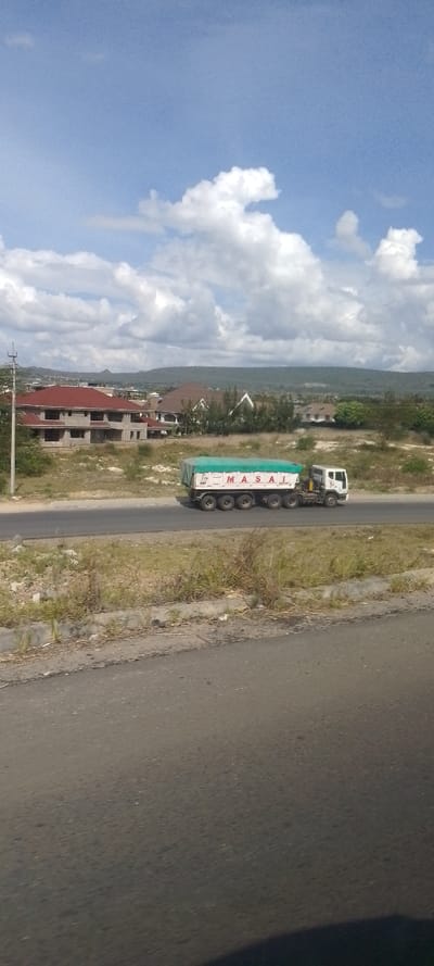 The image presents a semi-urban landscape in Kinanie ward, Kenya, under a daytime sky with scattered cumulus clouds. A white commercial articulated lorry, featuring a white cab and a multi-axle trailer covered by a green tarp, is traveling from left to right on a paved two-lane road. The word "MASAI" is clearly visible in red letters on the side of the trailer. Adjacent to the road, a strip of dry, sparse grass and scrubland separates it from the foreground, which consists of a dark asphalt surface with a dark shadow partially obscuring the lower right corner. A concrete curb marks the edge of this foreground section. In the background, several residential buildings are visible, some with red or brown tile roofs, while others appear to be under construction with exposed concrete walls. These structures are interspersed with trees and undeveloped land, leading to distant, vegetated hills under the horizon. An unidentified utility pole stands near the road on the left. No individuals are visible.