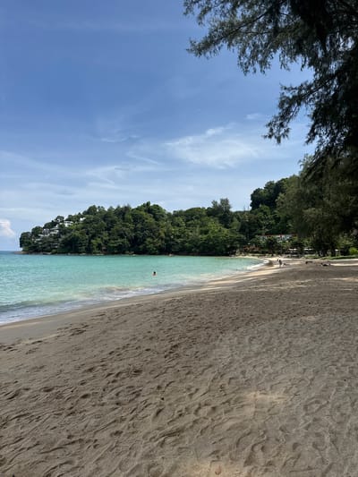 The image depicts a tropical beach scene in Ban Bang Thao, Thailand. The foreground is dominated by textured sand, showing numerous footprints. A gentle wave laps at the shore, separating the sand from the clear, turquoise ocean. In the mid-ground, the ocean extends towards a lush, green hillside covered in dense foliage and dotted with white buildings, suggesting a resort or residential area. A person can be seen swimming in the water. The background features a clear blue sky with scattered wispy clouds. To the right of the frame, the dark branches of pine trees overhang the scene, casting shadows on the sand. Several other people are visible on the beach and in the shallow water further down the coastline. The overall impression is one of a serene and picturesque beach environment.
